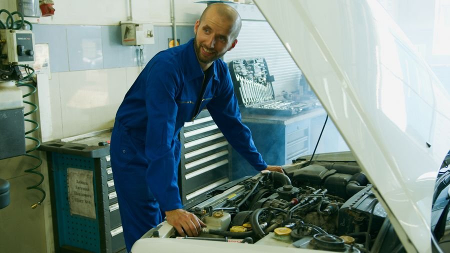 UK car mechanic in blue overalls smiling while inspecting an engine bay in a garage