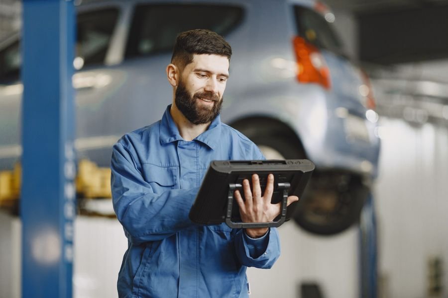 UK mechanic in blue overalls using a diagnostic tablet beside a car on a garage ramp