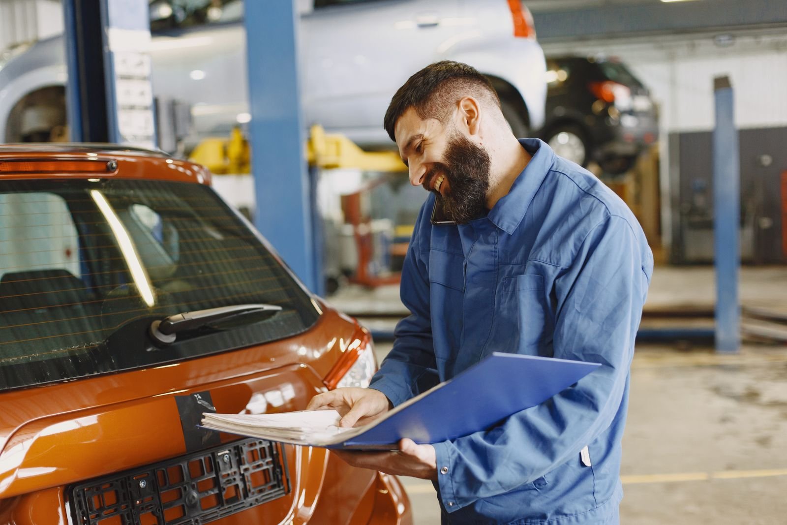 UK car mechanic in blue overalls smiling with a job sheet beside a car in a service garage