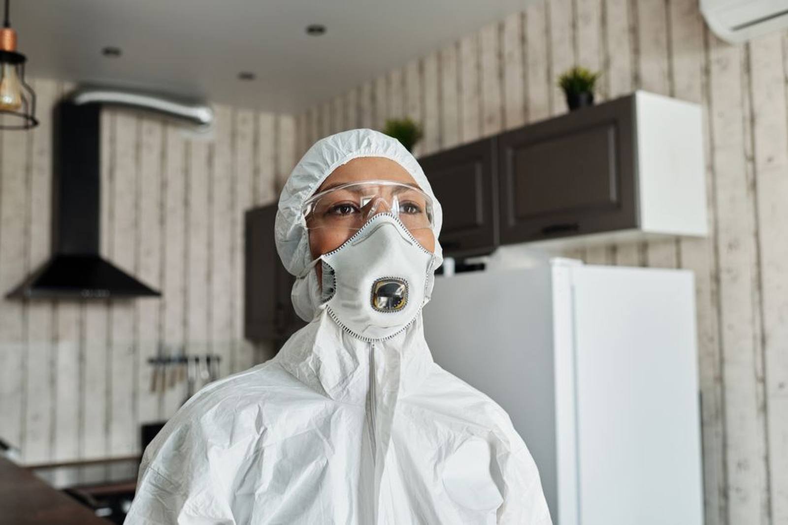 Pest control technician in protective coveralls and respirator inspecting a home kitchen
